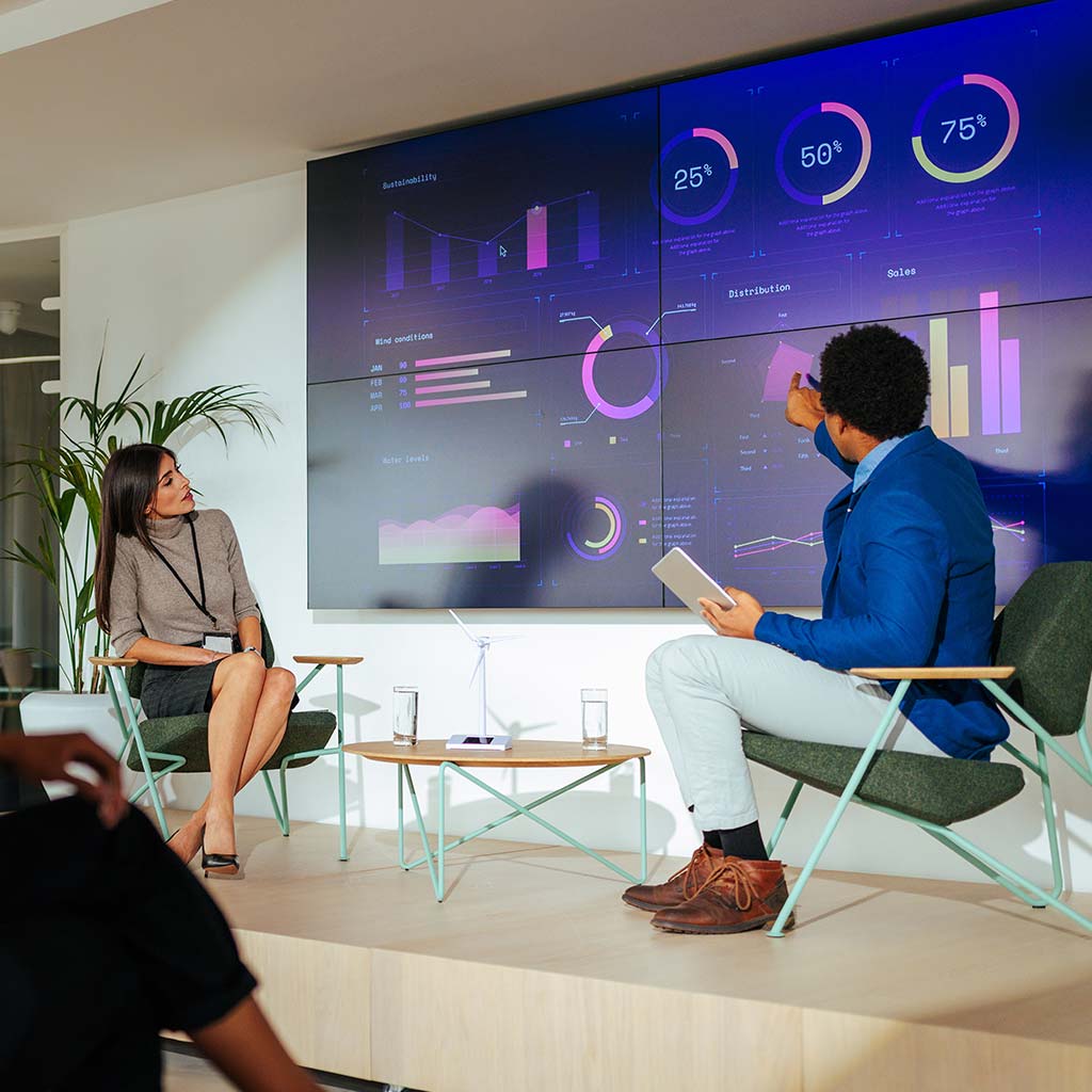 presenters on stage in front of large video wall containing graphs and charts
